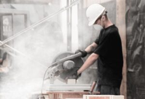 A worker in a hard hat and mask operates a circular saw to cut bricks, surrounded by dust in an active work environment.